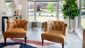 A room with two plush armchairs, a large mirror, a lamp, and a view of a backyard through large windows.
