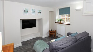 Ein modernes Wohnzimmer mit grauem Sofa, einem flachbildfernseher an der Wand, einem kleinen Holztisch und einem Fenster mit Blick nach draußen.