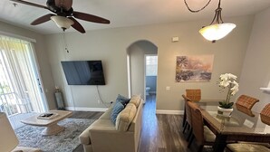 A modern living room with a sofa, coffee table, and dining table. A ceiling fan and TV are mounted on the wall. - Windsor Palms 3 bed ground floor condo near DISNEY! (Kissimmee)