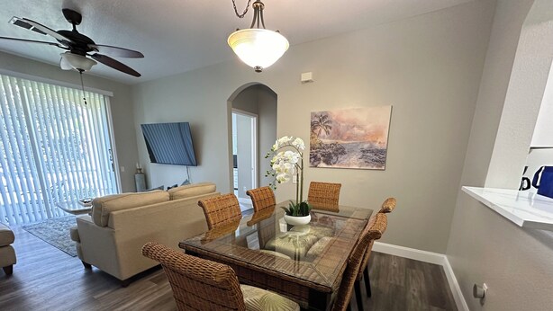 A dining area with a glass-top table, wicker chairs, a white vase with flowers, and a painting on the wall. - Windsor Palms 3 bed ground floor condo near DISNEY! (Kissimmee)