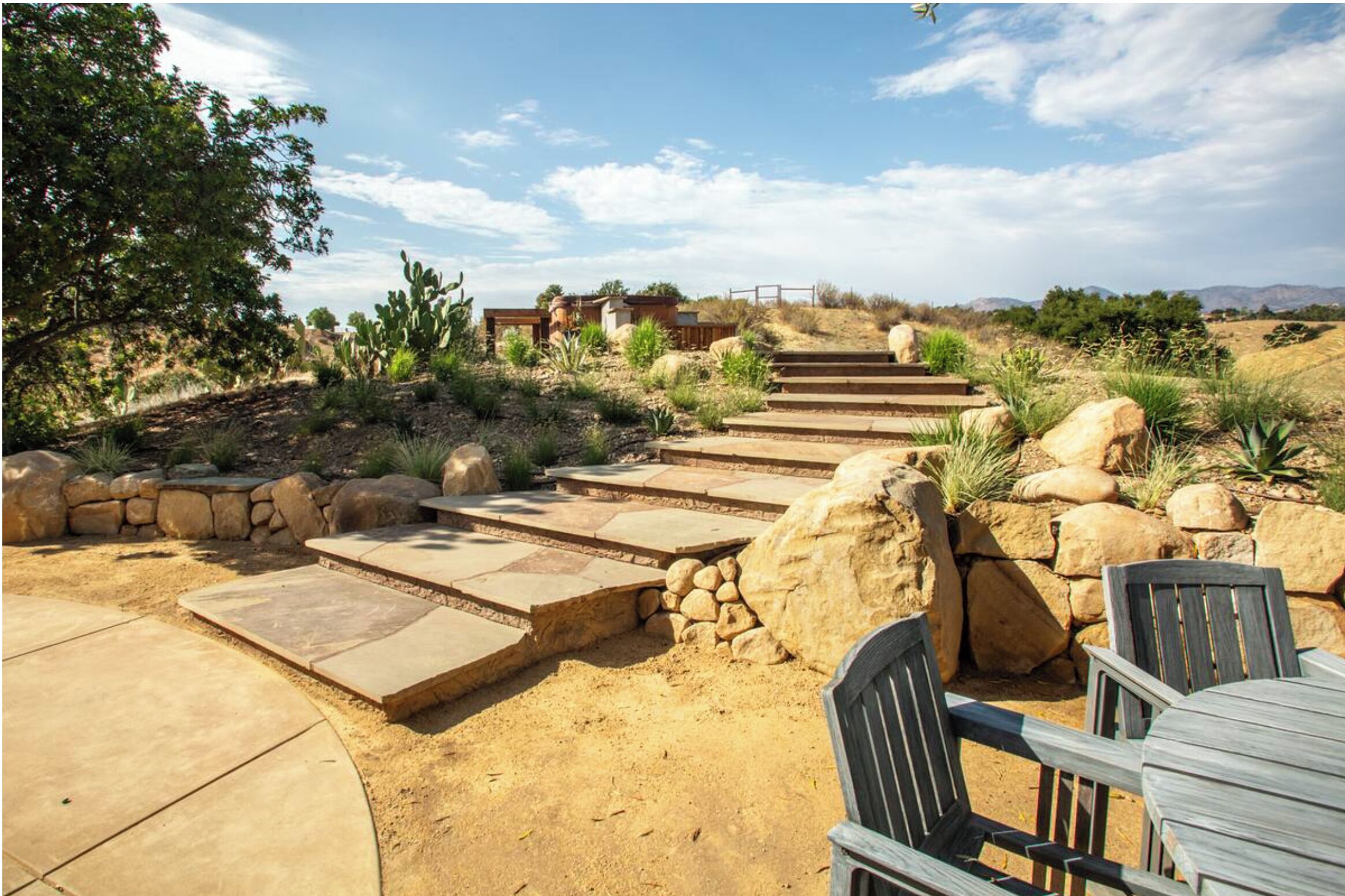 A patio area with stone steps leading to a wooden deck, surrounded by desert vegetation and a view of distant hills.