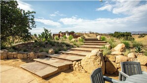 Un patio con escalones de piedra que llevan a una terraza de madera, rodeado de vegetación desértica y vistas a colinas distantes.