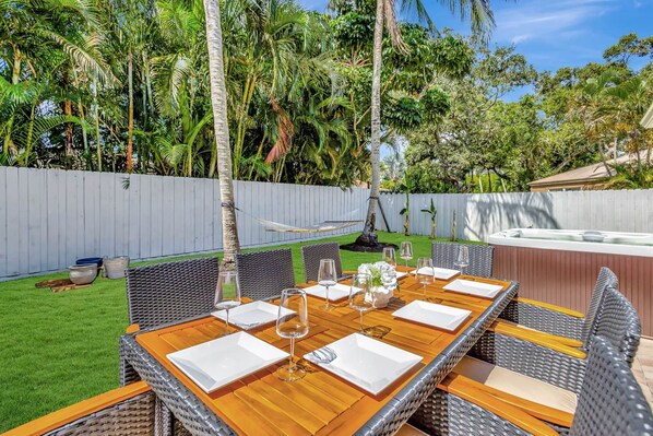 Un espacio de comedor al aire libre con una mesa de madera y sillas de mimbre, rodeado de exuberante vegetación y una cerca blanca.