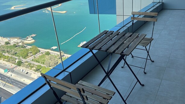 Ein Balkon mit Blick auf eine Küstenstadt, ausgestattet mit einem Glasgeländer und zwei hölzernen Bänken.