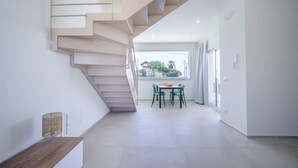 Un intérieur moderne avec un escalier menant à un balcon, un coin repas avec une table et des chaises, et une vue sur la verdure à l’extérieur.