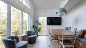 Moderni ruokailutila, jossa on puinen pöytä ja tuolit, suuret ikkunat, joista avautuu näkymä puita kohti, sekä seinälle asennettu televisio.