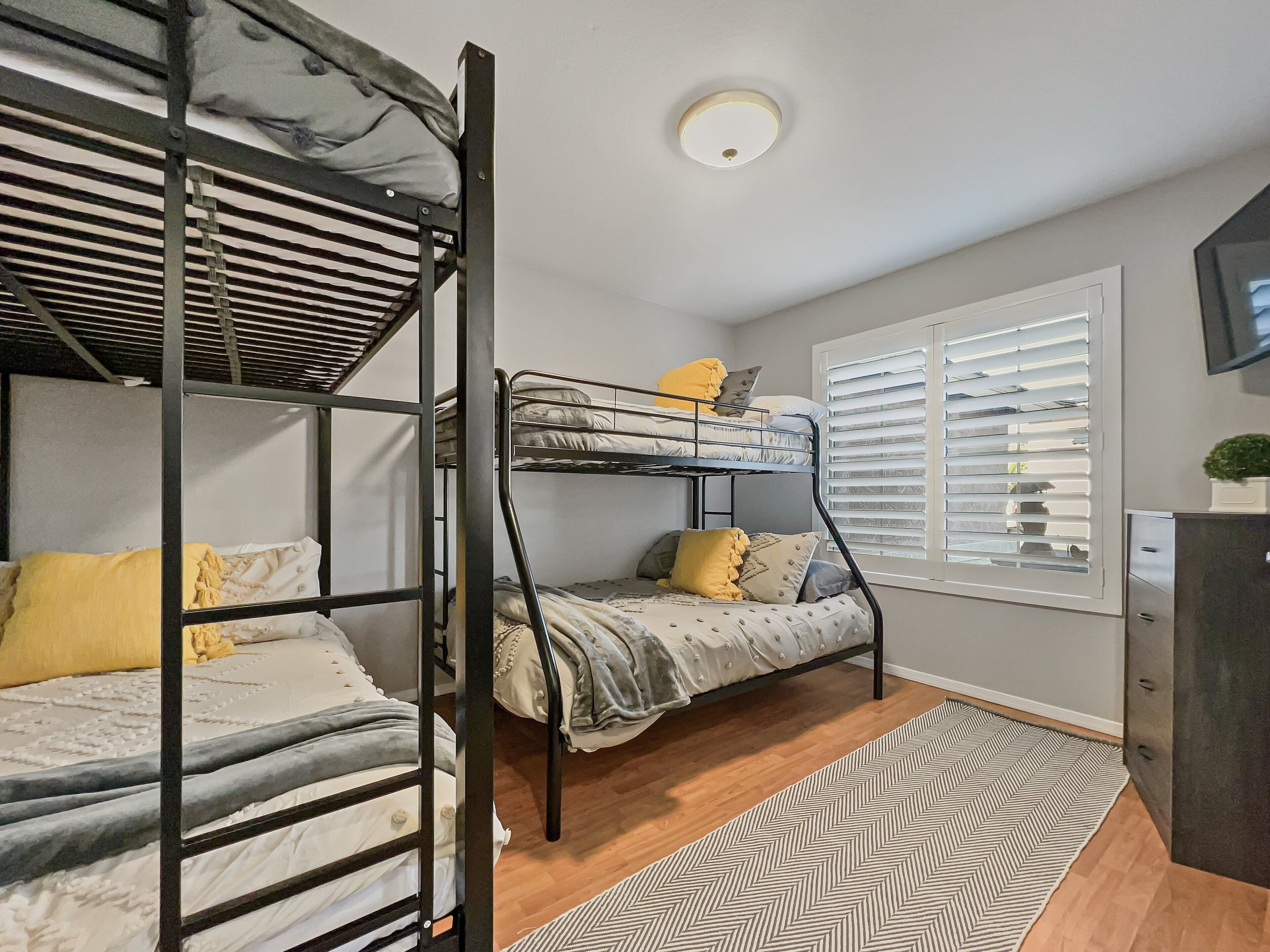 A bunk bed room with a herringbone-patterned rug, a desk with a plant, and a window with blinds.