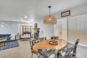 Uma sala de estar espaçosa com um sofá azul, uma mesa de jantar redonda de madeira, cadeiras estampadas, um lustre de vime e vista através de grandes janelas com persianas.