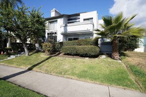 Um edifício residencial moderno de dois andares com telhado plano, janelas amplas e um gramado bem cuidado com uma palmeira.