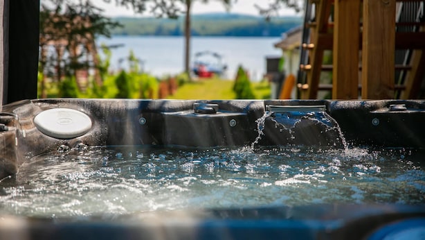 A hot tub with water splashing, surrounded by greenery and a view of a lake. - La Vue O-Lac Magog (Sherbrooke)