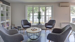 Ein modernes Wohnzimmer mit einem runden Couchtisch, zwei Sesseln, einem Bücherregal und Blick auf einen See durch große Fenster.