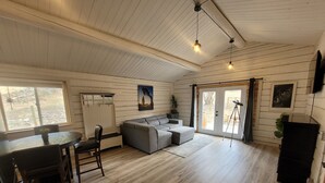 A spacious living area with a wooden floor, a grey sectional sofa, a dining table with chairs, a small kitchen area, and a sliding glass door leading outside.