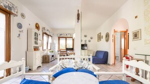 Un salón acogedor con un sofá blanco, una mesa redonda y una silla azul. Hay una zona de cocina con una estufa y otra silla azul. El ambiente tiene suelo de baldosas y paredes blancas decoradas con platos.