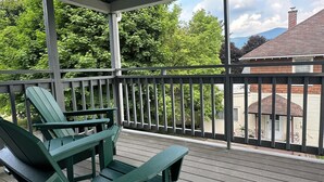 A covered porch with green Adirondack chairs, a wooden deck, and a view of a house and trees. - Spacious ski chalet with pool table & mountain views (Rutland)