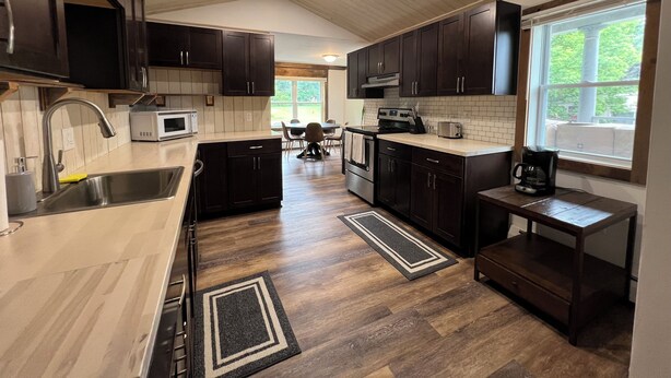A modern kitchen with dark wooden cabinets, a stainless steel sink, a microwave, and a large window providing natural light. - Spacious ski chalet with pool table & mountain views (Rutland)