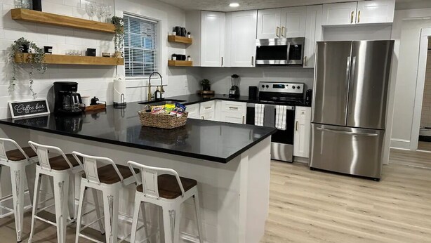 Una cocina moderna con gabinetes blancos, electrodomésticos de acero inoxidable, una isla central con taburetes y una zona de café.