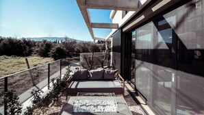 A modern outdoor seating area with a cushioned bench and a glass table, overlooking a natural landscape. - Terra Segreda (Volos)