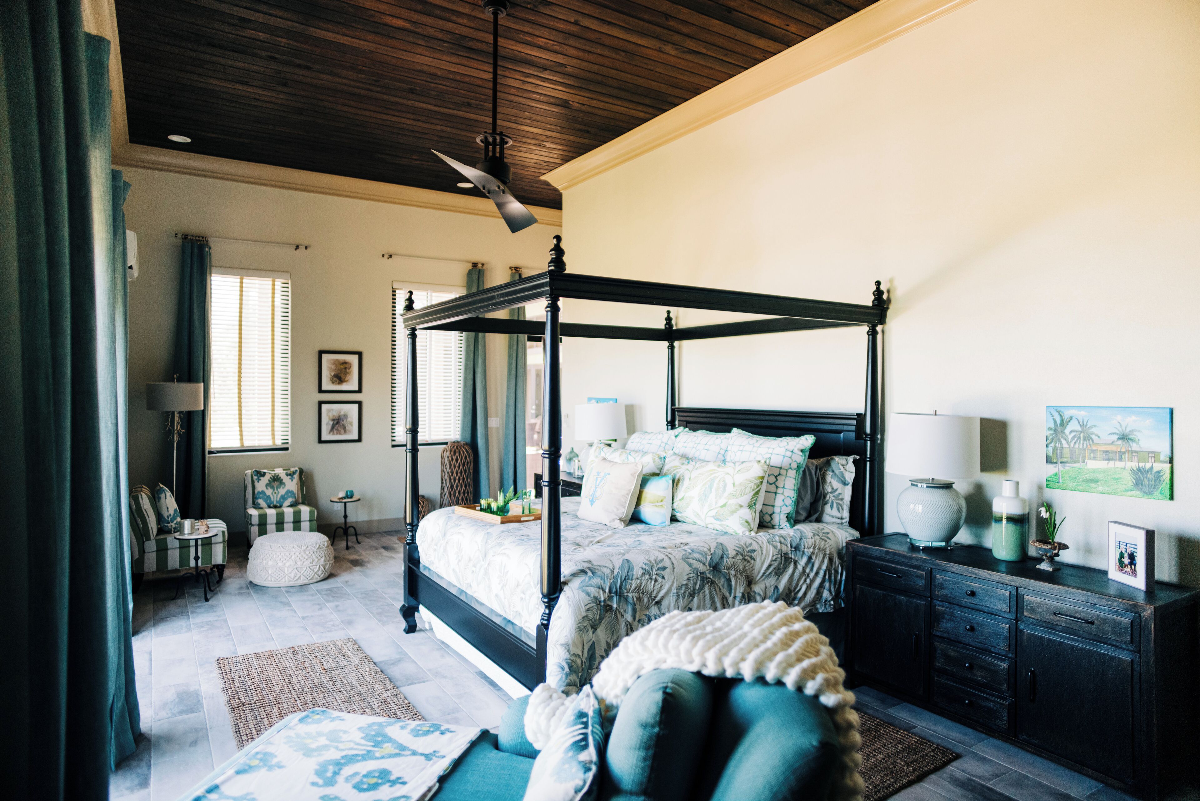 A spacious bedroom with a four-poster bed, a sitting area with a chair and ottoman, a small table, and a dresser with a mirror.