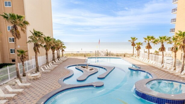A swimming pool with a wavy design, surrounded by lounge chairs and palm trees, with a beach and ocean view in the background. - Crystal Tower Corner w/ 2 Balconies, 3 Pools, Lazy River, Free Daily Activities (Gulf Shores)