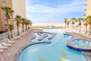 A swimming pool with a wavy design, surrounded by lounge chairs and palm trees, with a beach and ocean view in the background. - Crystal Tower Corner w/ 2 Balconies, 3 Pools, Lazy River (Gulf Shores)