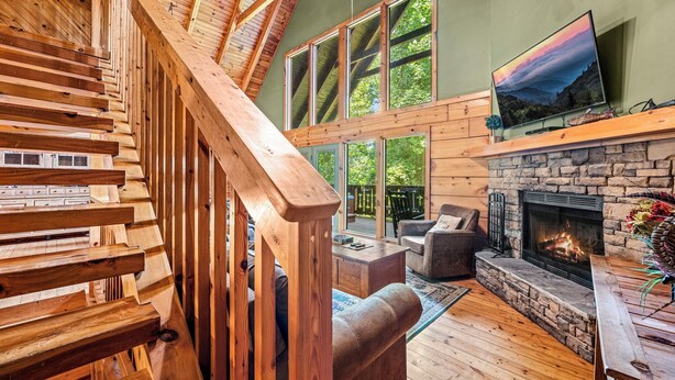 A spacious living area with a stone fireplace, wooden staircase, and large windows overlooking a deck. - Spring With A View! Hot Tub, Theater, Pool Table (Gatlinburg)