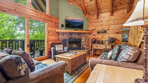 A cozy cabin interior with a stone fireplace, wooden furniture, and large windows overlooking a forest. - Spring With A View! Hot Tub, Theater, Pool Table (Gatlinburg)
