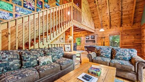 A log cabin interior with a wooden staircase, two patterned sofas, a wooden coffee table, and a view of the outdoors through a window. - Spring With A View! Hot Tub, Theater, Pool Table (Gatlinburg)
