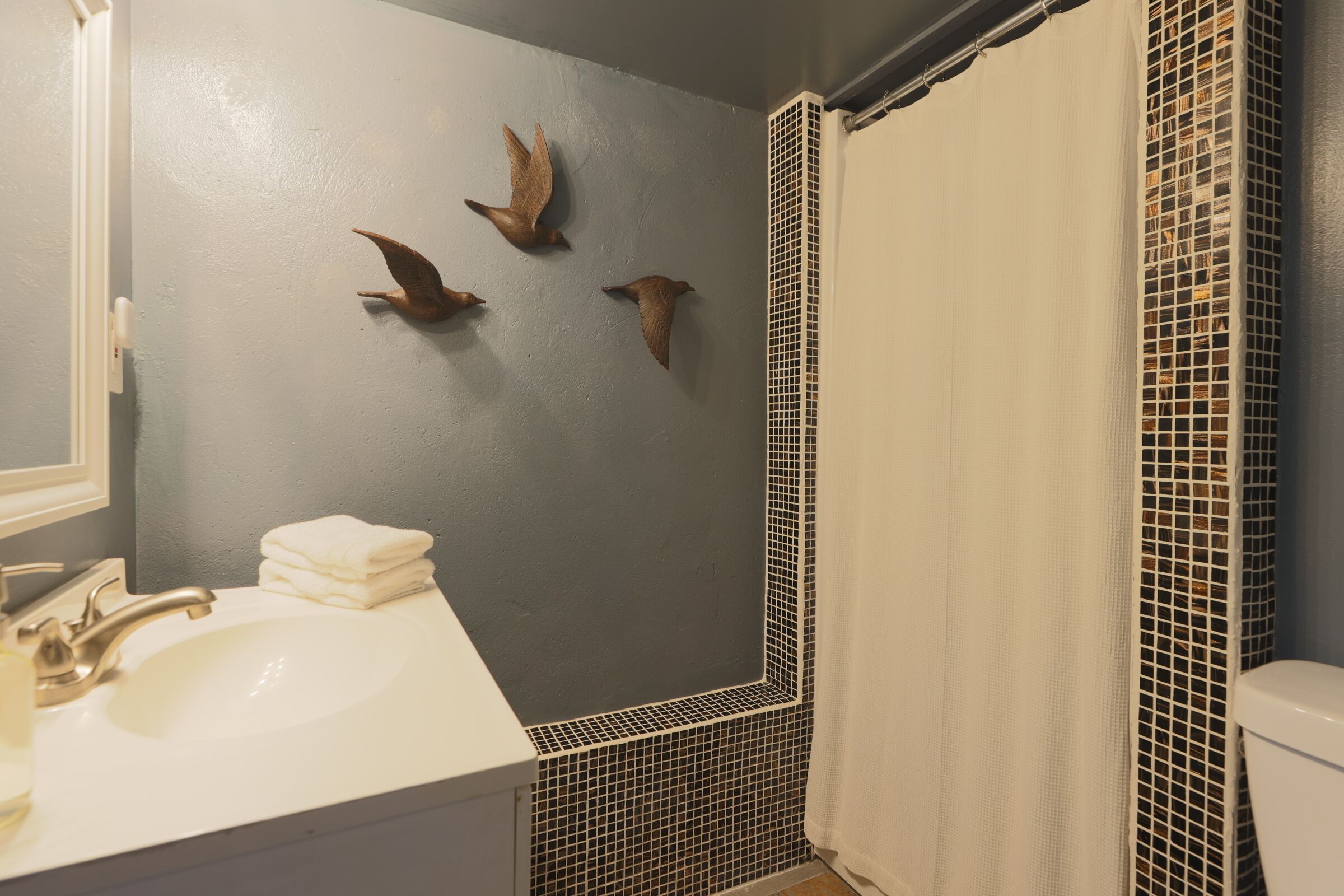Un baño con lavamanos, espejo y tres adornos de pájaros colgados en la pared.