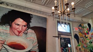 A person holding a bowl of soup in a room with a decorative ceiling, a wall with a colorful mural, and an exit sign. - 4bed studio basement! 5min->downtownKC (Kansas City)