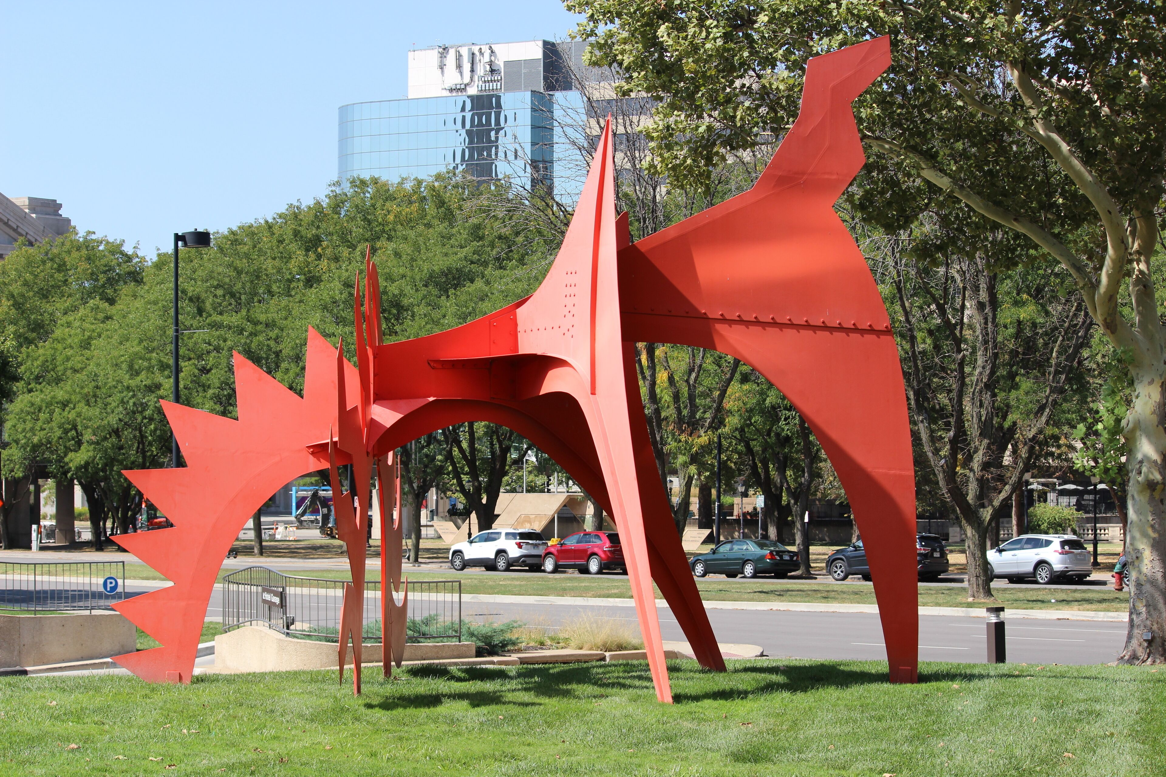 Eine rote, abstrakte Skulptur in einem Park mit Bäumen und einem Parkplatz im Hintergrund.