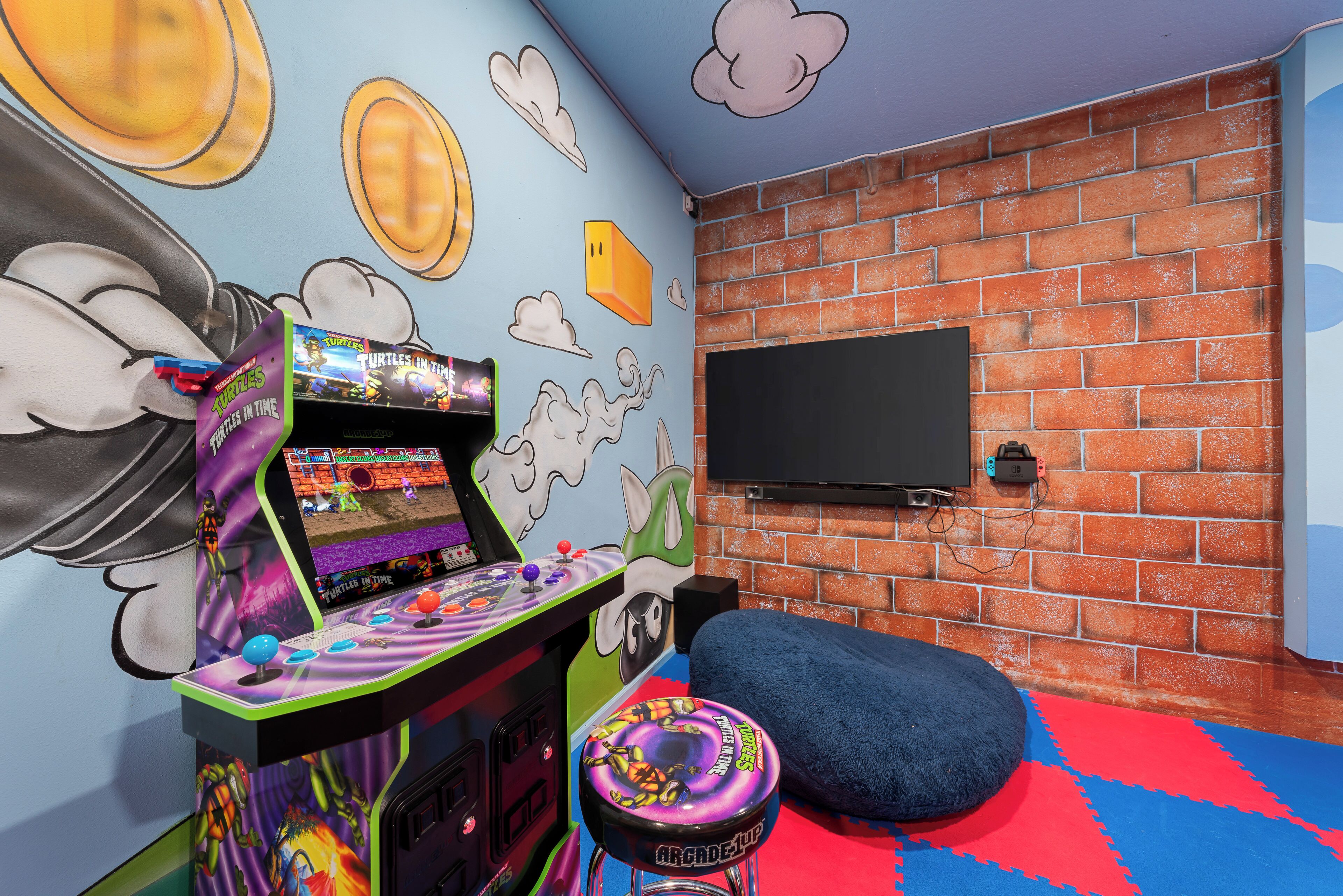 Une pièce avec un mur en briques, un téléviseur à écran plat fixé au mur, une borne d’arcade colorée et un fauteuil en forme de sac de haricots bleu, posé sur un tapis multicolore.