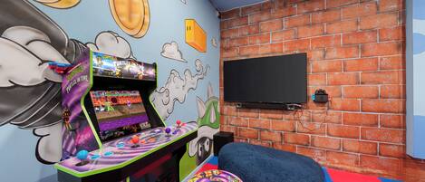 Une pièce avec un mur en briques, un téléviseur à écran plat fixé au mur, une borne d’arcade colorée et un fauteuil en forme de sac de haricots bleu, posé sur un tapis multicolore.