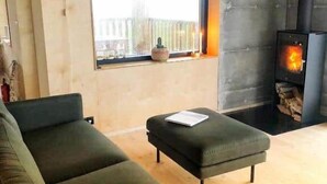 A modern living room with a sofa, a small ottoman, a fireplace, and a window with a view of the outside. - Modern mountain-view cottage in Golden Circle with outdoor Hot Tub & Sauna (Hrunamannahreppur)