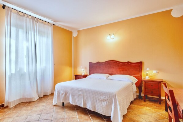 A bedroom with a large bed, two bedside tables with lamps, a wooden headboard, and a window with sheer curtains.
