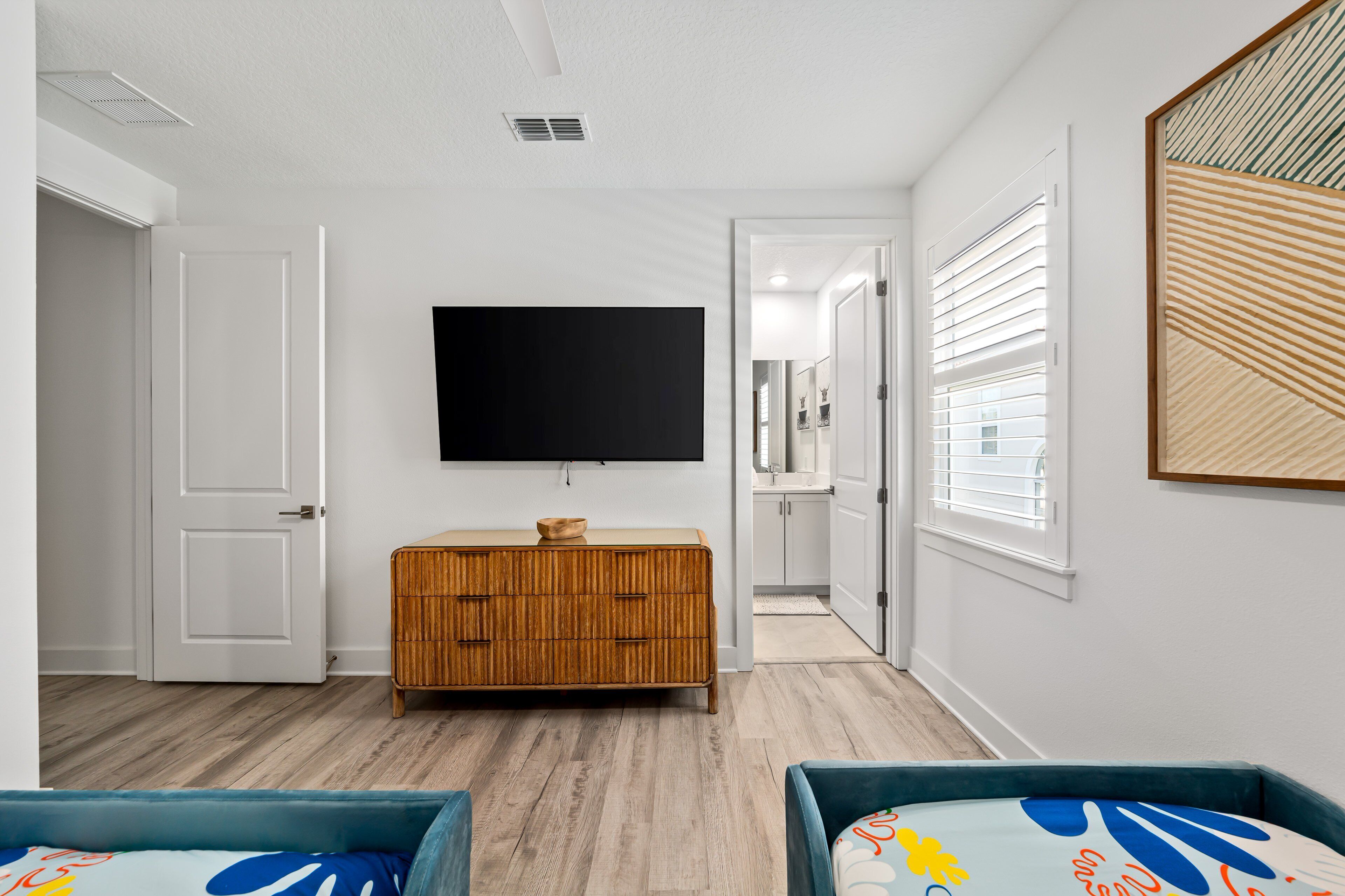 A modern bedroom with a wooden dresser, a flat-screen TV mounted on the wall, and a bed with a colorful headboard.