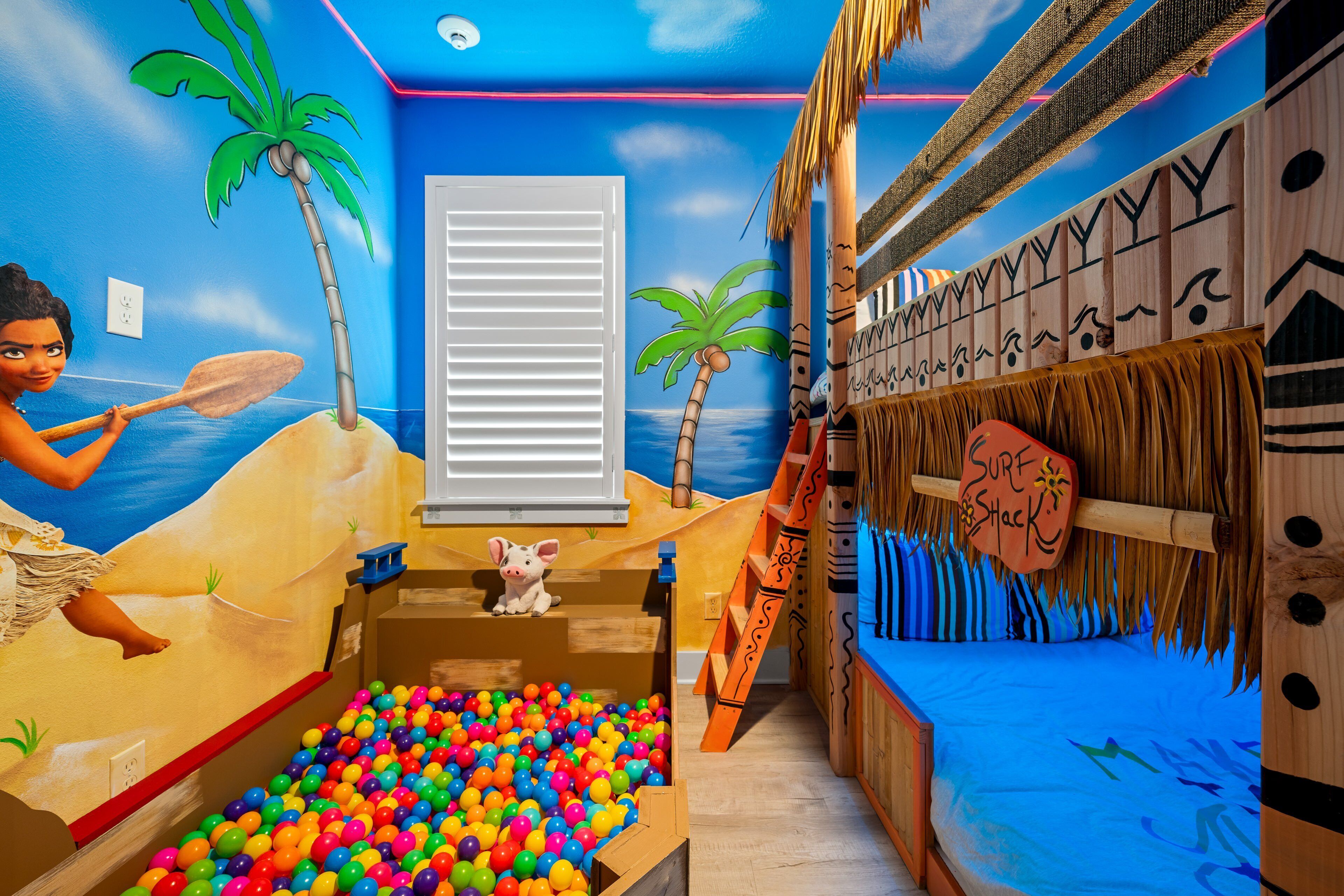 A room with a beach-themed mural, a ball pit, a bed with a 'Sure Shack' sign, and a ladder leading to a platform.