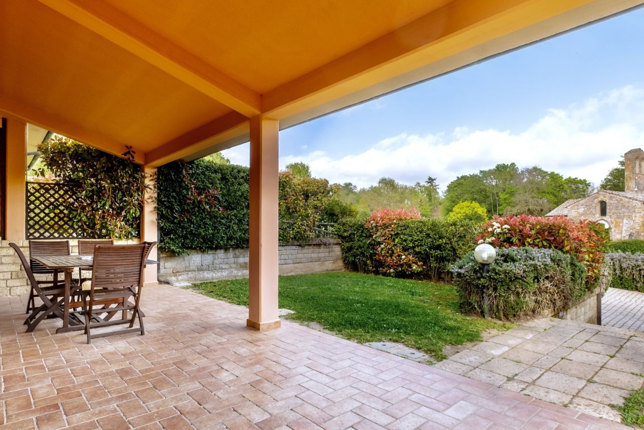 A covered patio with a brick floor, a wooden table and chairs, and a view of a well-maintained garden with various plants and a stone building in the background.