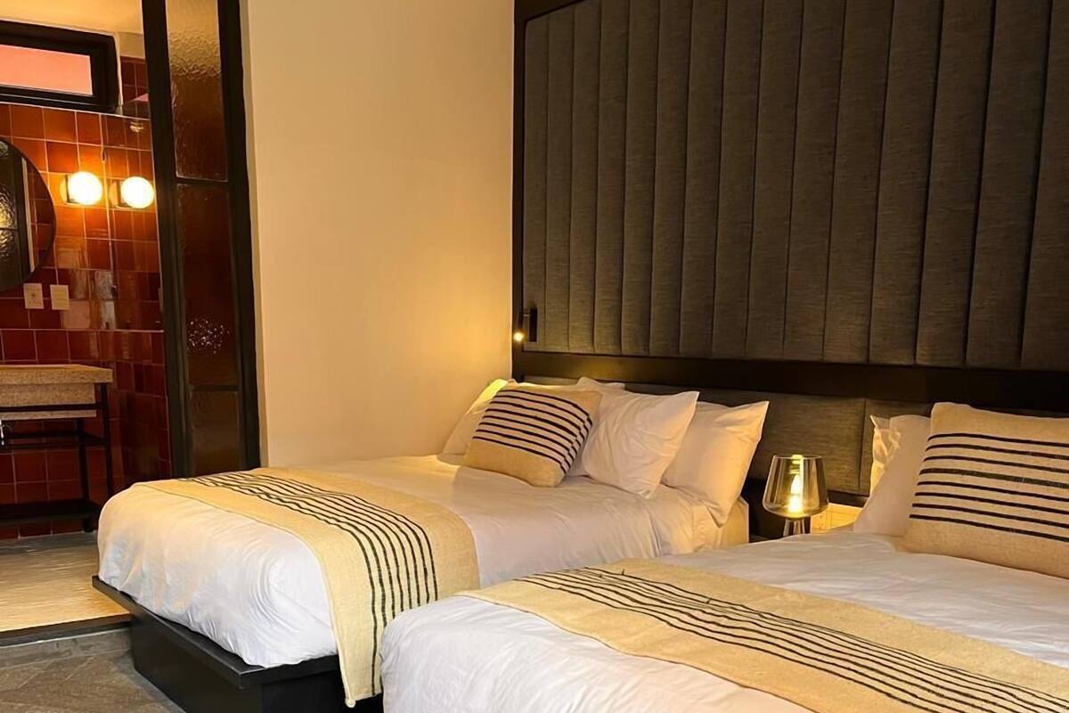 Une chambre d’hôtel avec deux lits, une grande tête de lit rembourrée et une salle de bain visible à travers une porte ouverte.