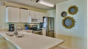 A kitchen with white cabinets, a stainless steel refrigerator, a microwave, and a coffee maker. There are three sunflowers on the wall. - 🦩Book Me Baby! Bodacious Beach Retreat Condo in Destin🦩 (Destin)