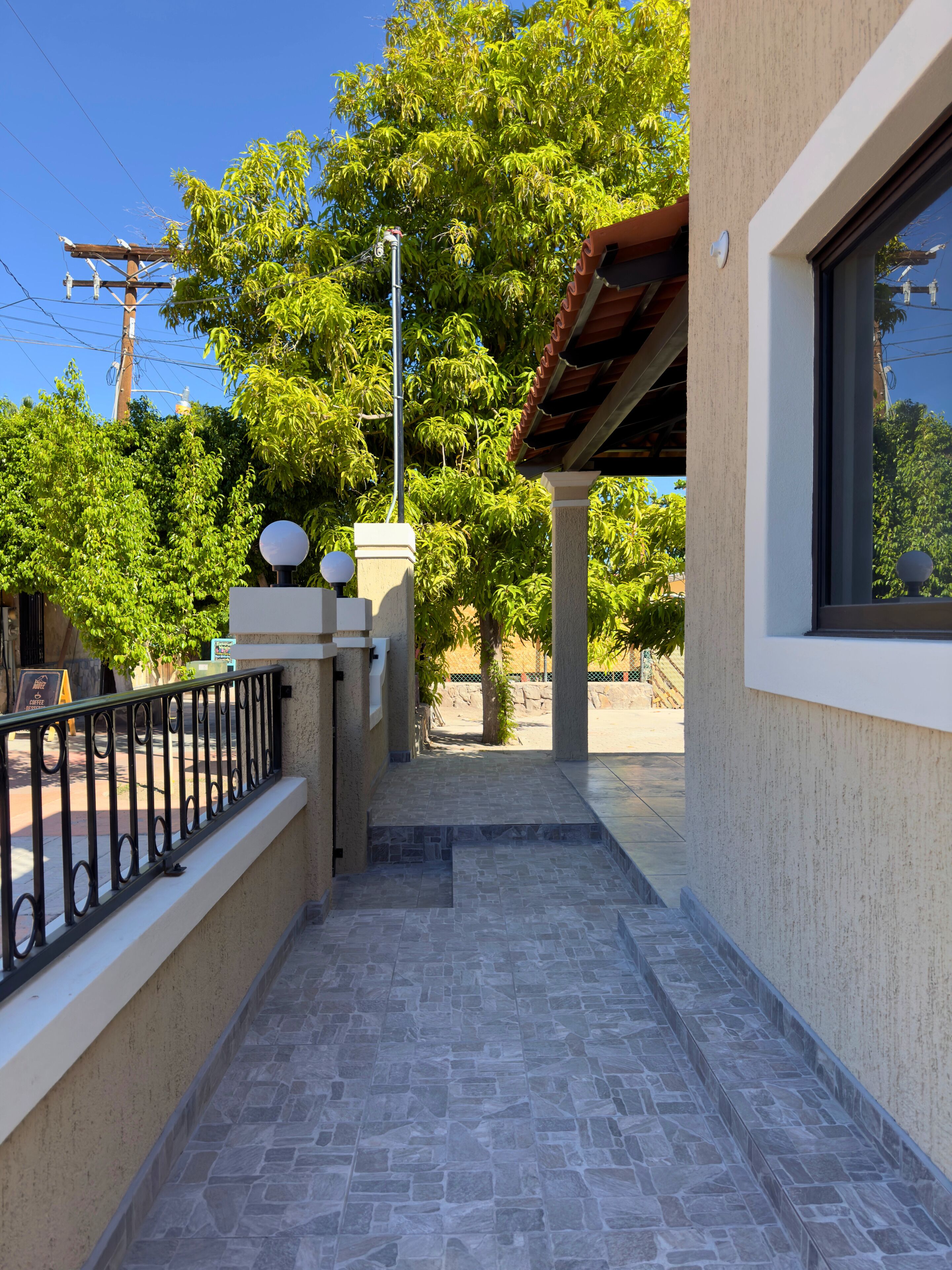 A narrow outdoor corridor with a stone floor, a metal railing, and a view of a tree and a building in the background.
