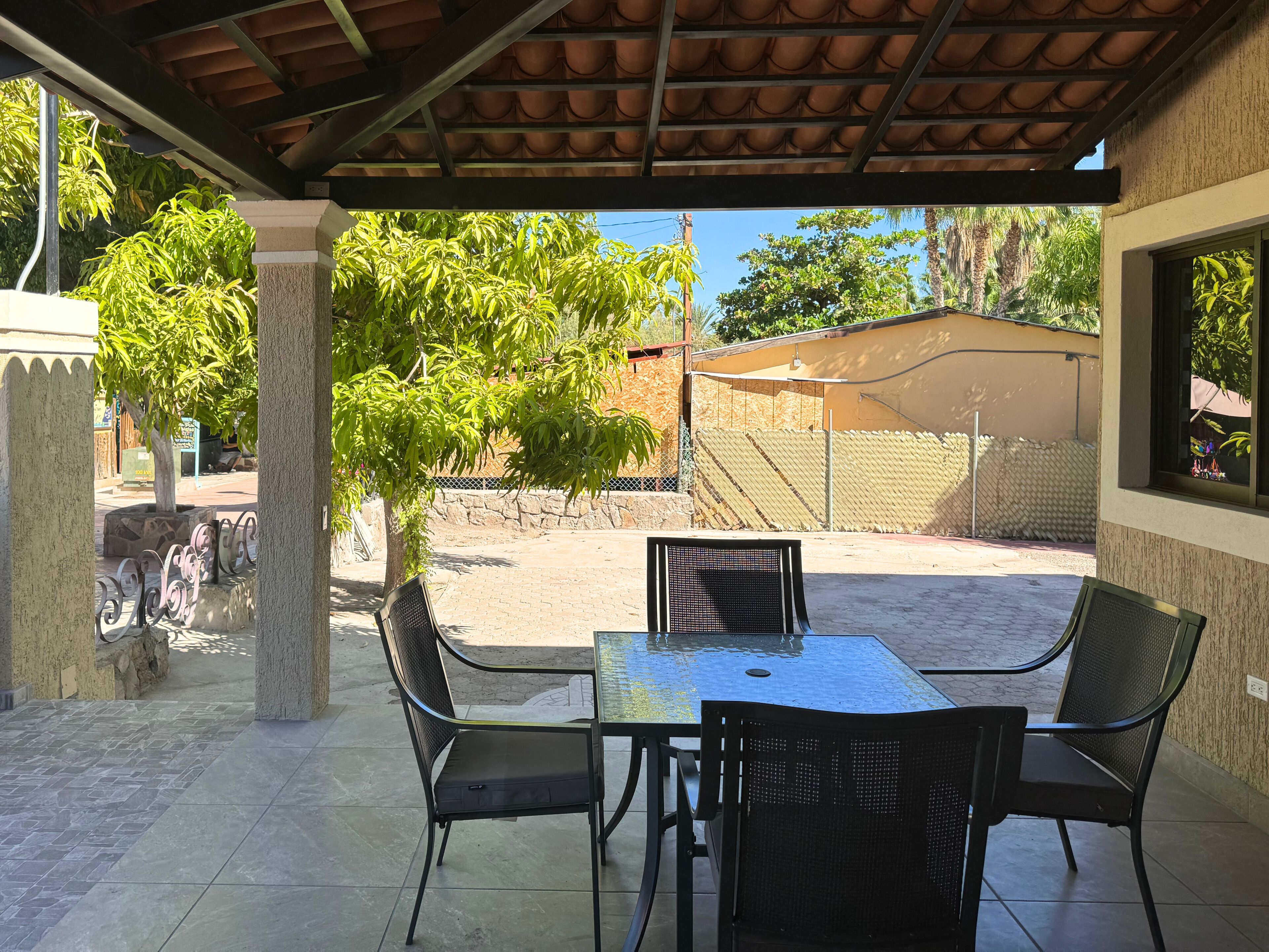 Alpendre coberto com mesa e cadeiras, uma árvore e vista para uma área cercada com um edifício ao fundo.