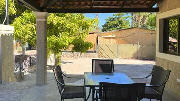 Un patio cubierto con una mesa y sillas, un árbol y vistas a un área cercada con un edificio al fondo.
