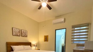 A bedroom with a bed, a ceiling fan, a nightstand, a lamp, a wall-mounted air conditioner, and a window with blinds. - Posada Don Esteban by PartnerHost (Loreto)