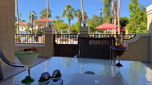 A glass table with a martini glass and a bowl of fruit, a laptop, and sunglasses on a balcony with a view of palm trees and a gate. - Posada Don Esteban by PartnerHost (Loreto)