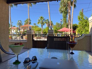 Een glazen tafel met een martini glas en een schaal fruit, een laptop en zonnebril op een balkon met uitzicht op palmstruiken en een poort.