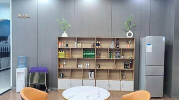 A room with a marble table, two orange chairs, a bookshelf with various items, and a grey refrigerator.