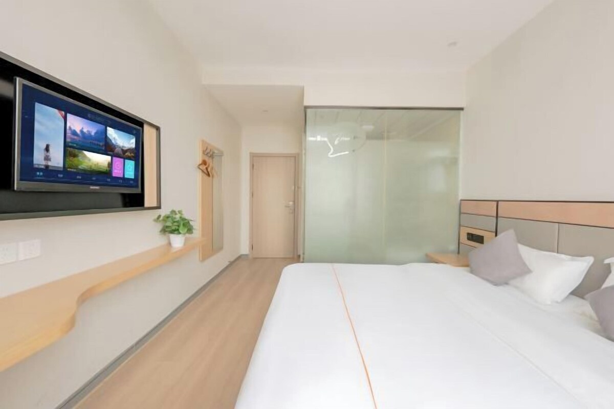 A modern hotel room with a large flat-screen TV mounted on the wall, a bed with white bedding, a small potted plant, and a sliding glass door leading to a shower area.