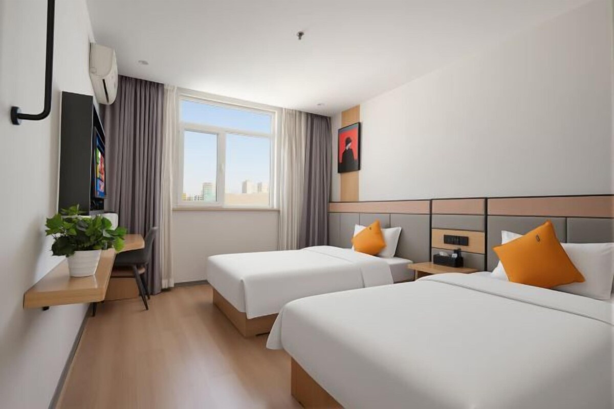 A hotel room with two beds, a desk, a chair, a plant, and a window with curtains.