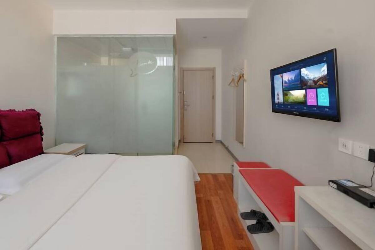 Una habitación de hotel moderna con una cama, un televisor montado en la pared, un banco rojo y una puerta de vidrio que da al baño.