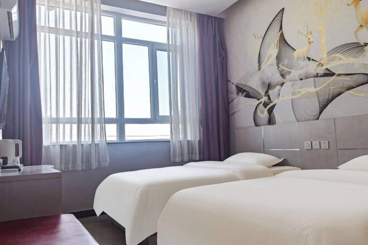 Habitación de hotel con dos camas, un ventanal con cortinas translúcidas, un aire acondicionado de pared y un cuadro con formas abstractas.